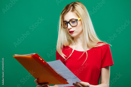 Smart Female Student in Eyeglasses and Red Top is Reading Something on Green Background in Studio.