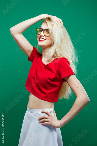 Fashionable Amazing Blonde Model with Long Hair and Eyeglasses Wearing White Skirt and Short Red Top is Posing in Studio on Green Background.