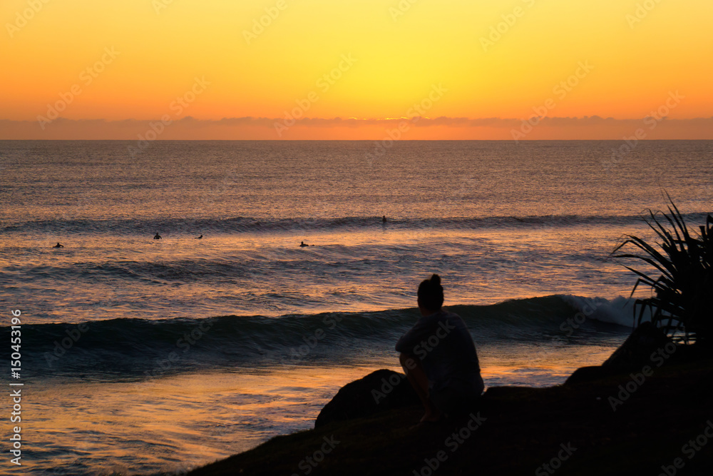A Silhouetted Girl Sits on Rocks Watching Sunrise Surfers