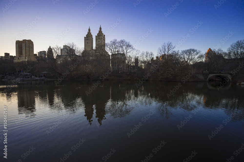 Scenic sunset view of the New York City skyline with bare winter trees reflecting with buildings of the Upper West Side in the Central Park lake
