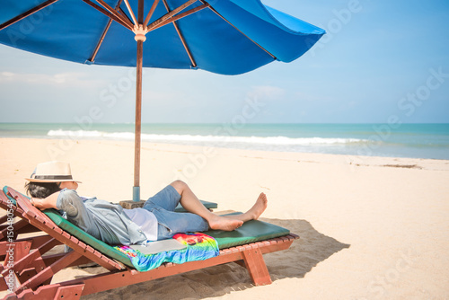 young man lying on wooden beach bench with blue umbrella and close his face by hat, beautiful tropical beach island and blue sky, vacation time and summer holiday concepts, digital nomad lifestyle © zephyr_p