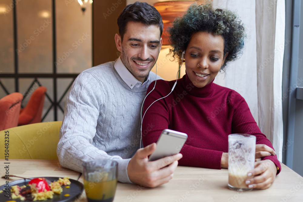 Loving mixed couple staying in cafe, European male hugging his ethnic girlfriend, they are both listening to audio tracks from his smartphone sharing earphones, smiling, looking happy and pleased. Loving mixed couple staying in cafe, European male hugging his ethnic girlfriend, they are both listening to audio tracks from his smartphone sharing earphones, smiling, looking happy and pleased.