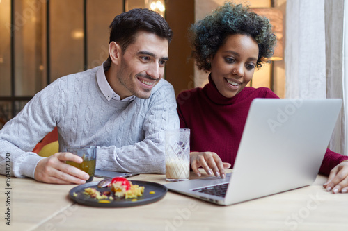 Closeup of young interracial lovers spending time in cafe watching media together on laptop. African American lady is using touchpad, Caucasian male is looking at display with surprise holding teacup.