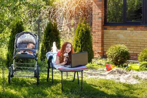 Young woman with her newborn baby lying on the soft armchair with laptop. Summer day. Mother with child outdoors. Motherhood.