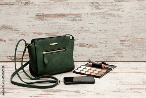 Green ladies handbag, phone, eyeshadow palette and a lipstick on a wooden background. fashion concept photo