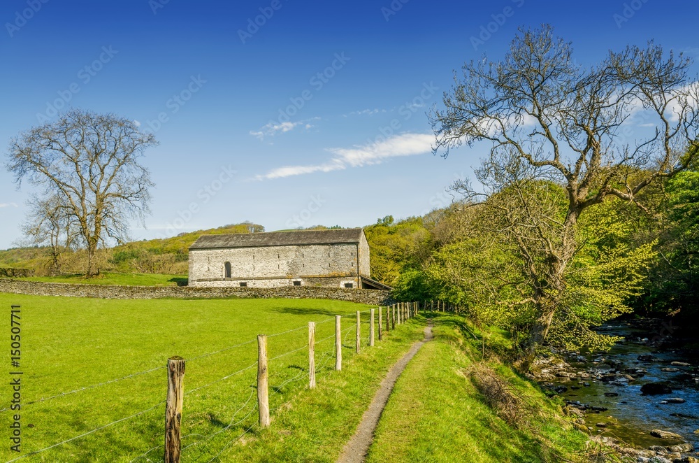 An isolated barn set in green English countyside by a path running alongside a river.
