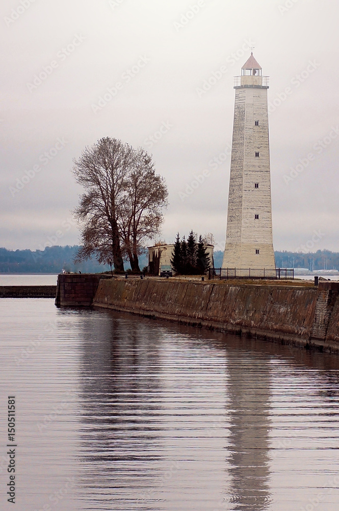 Tower lighthouse stands on the bank of a canal next to a tree, reflected in water