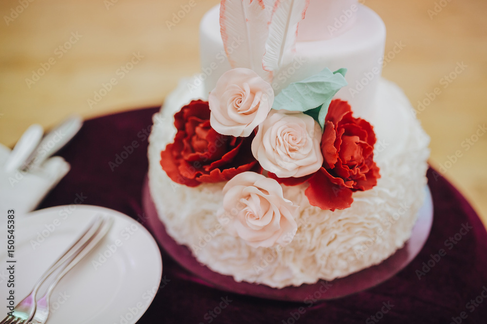 Three-tiered wedding cake is decorated with flowers and stands on the table next to a plate and cutlery