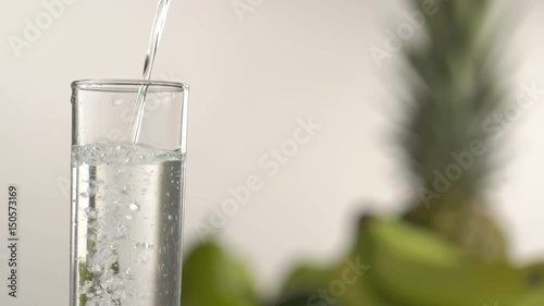 The close-up view of the water pouring into the high glass at the blurred background of the green apples, pineapples and pears. photo