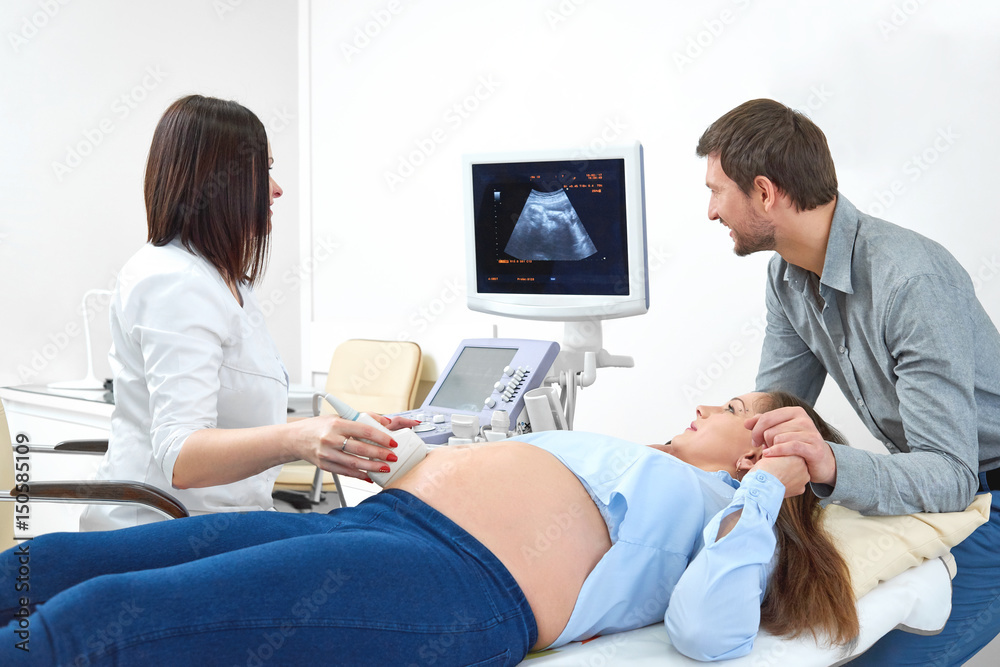 Obraz premium Pregnant young woman holding hand of her loving husband during medical examination at the clinic. Female doctor performing ultrasound scanning on her pregnant patient pregnancy fertility expecting.