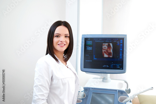 Young female doctor smiling happily to the camera posing at her office near ultrasound scanning machine copyspace medical industry technology modern equipment diagnostics sonography healthcare.