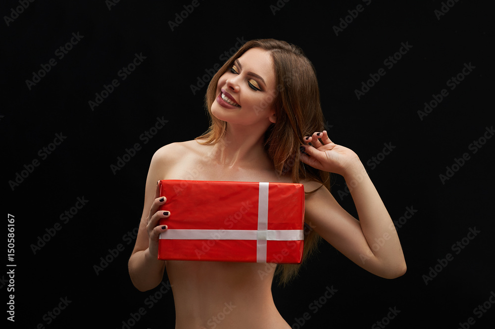 Gorgeous cheerful young woman posing topless covering her breasts with a  gift box smiling joyfully with her eyes closed touching her hair  flirtatiously on black background sexuality seduction surprise Stock Photo