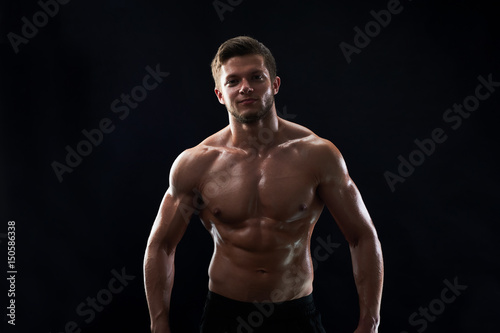 Handsome young shirtless athletic man smiling to the camera posing confidently on black background showing off his sexy strong body six-pack abs muscles fitness happiness lifestyle sports motivation.