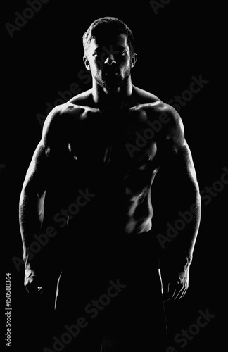 Vertical black and white silhouette of a shirtless athletic man with muscular strong body posing confidently on black background masculine brutal sportsman bodybuilder fitness athleticism concept.