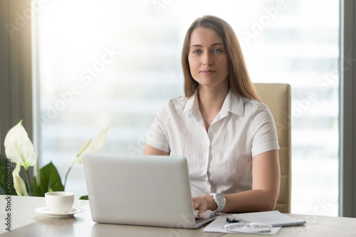 Portrait of confident businesswoman sitting at desk in office and working on laptop. Attractive woman typing on computer and looking at camera. Female office worker doing daily work with pleasure