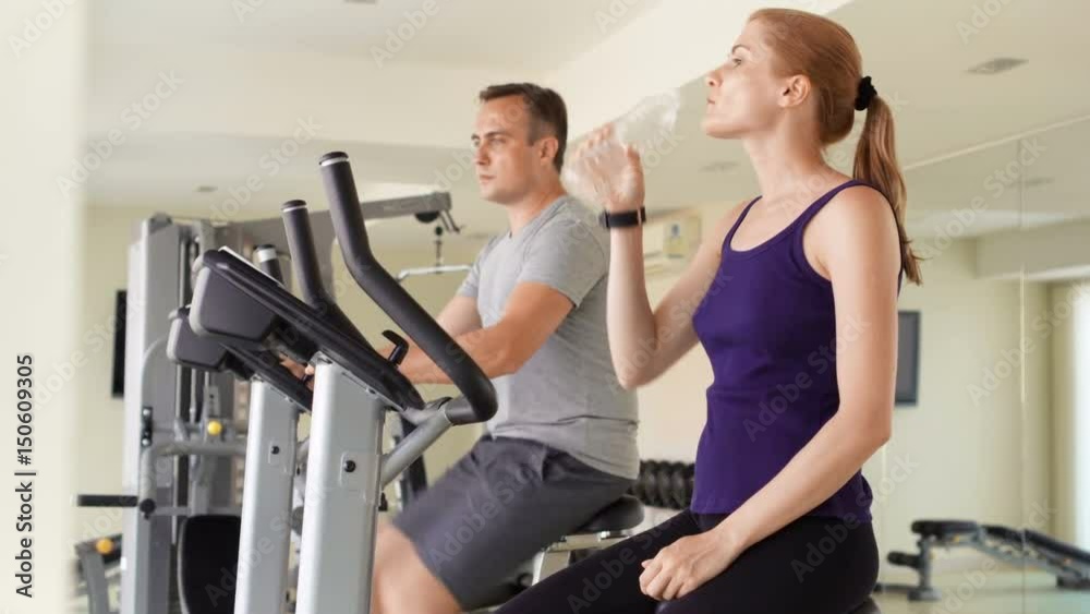 Fit sportive positive couple working out on exercise bikes at gym. Drinking water from plastic bottle Fit sportive positive couple working out on exercise bikes at gym. Drinking water from plastic bottle