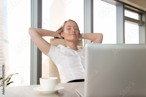 Businesswoman takes minute break for coffee and leaning back in chair with hands behind head. Successful female entrepreneur relaxing with satisfaction at workplace. Pleasure from great job done