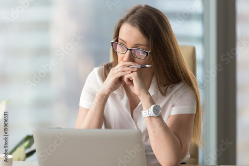 Portrait of pensive business woman seating at the table in office. Focused middle-aged woman in glasses resting her head on hands and thinking. Concentrated female looks aside and ponders decision