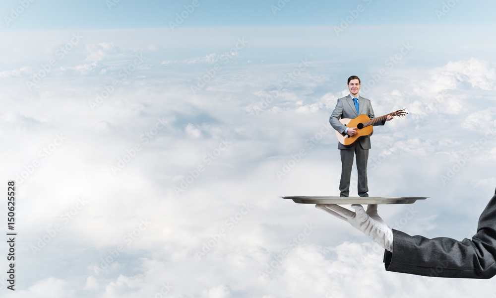 Businessman on metal tray playing acoustic guitar against blue sky background