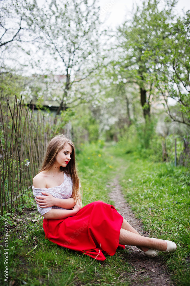 Portrait of sitting beautiful girl with red lips at spring blossom garden on green grass, wear on red dress and white blouse. Portrait of sitting beautiful girl with red lips at spring blossom garden on green grass, wear on red dress and white blouse.