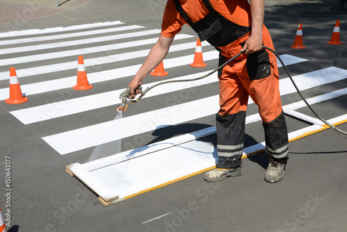 Worker is painting a pedestrian crosswalk. Technical road man worker painting and remarking pedestrian crossing lines on asphalt surface using paint sprayer gun.