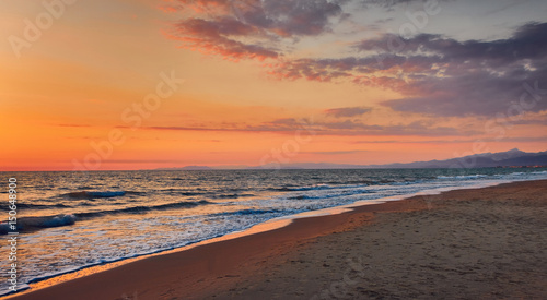 Panorama of a deserted beach at sunset. Turquoise foam waves on a sandy beach  golden sky with clouds from the setting sun  foggy blue silhouette of mountains in the background