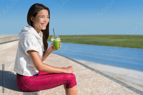 Healthy morning breakfast runner athlete drinking green smoothie juice drink cup before race for an active workout. Asian running woman happy looking at stadium blue tracks before cardio workout.