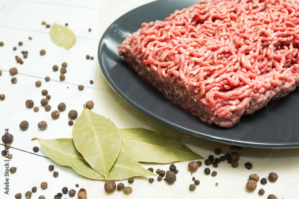 Portion of juicy fresh minced meat on a black plate on a wooden table with pepper and bay leaf.