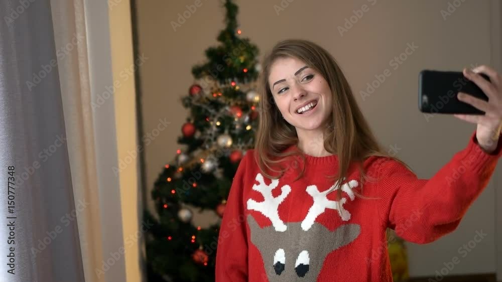 Young woman in sweater with geer taking selfie on smart phone on christmas tree background, she posing and smiling on camera. Young woman in sweater with geer taking selfie on smart phone on christmas tree background, she posing and smiling on camera.