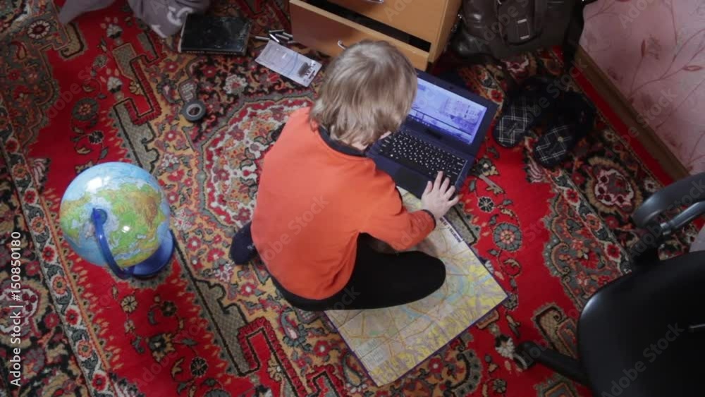 A little boy sitting on the floor in the children room at home and trying to do homework with a computer, maps, magnifier and globe. New generation, technology and new school education