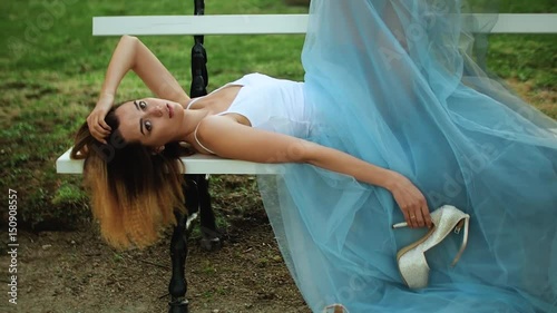 Attractive girl in white and blue dress lies on bench and handles high heeld shoes, talks, smiles while posing during photo shoot. photo