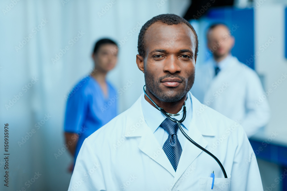portrait of confident doctor with stethoscope with colleagues behind in clinic portrait of confident doctor with stethoscope with colleagues behind in clinic