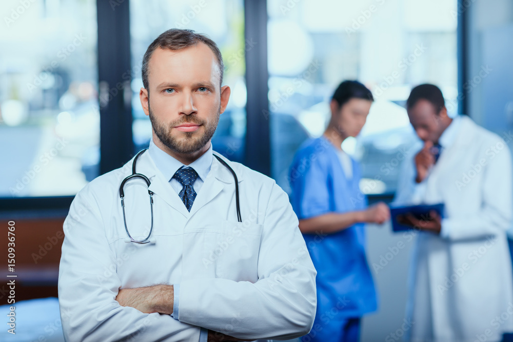 portrait of confident doctor with stethoscope with colleagues behind in clinic portrait of confident doctor with stethoscope with colleagues behind in clinic