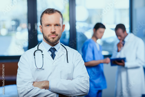 portrait of confident doctor with stethoscope with colleagues behind in clinic