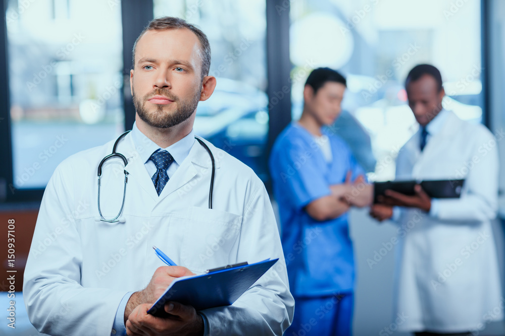 portrait of confident doctor writing in folder in clinic with colleagues behind portrait of confident doctor writing in folder in clinic with colleagues behind