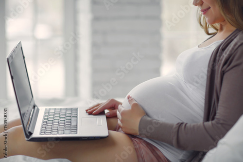 Portrait of a beautiful pregnant woman sitting on the bed with the computer. The concept of work during pregnancy. Side view.