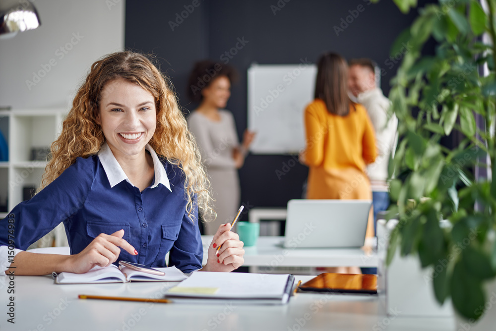 Young happy people discussing business standing at the whiteboard in modern light office, beautiful woman sitting at the table at forefront