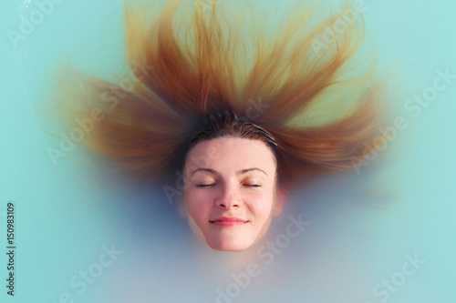 Woman with closed eyes floats in blue lagoon spa resort Blue Lagoon in Iceland, her red hair is around the head