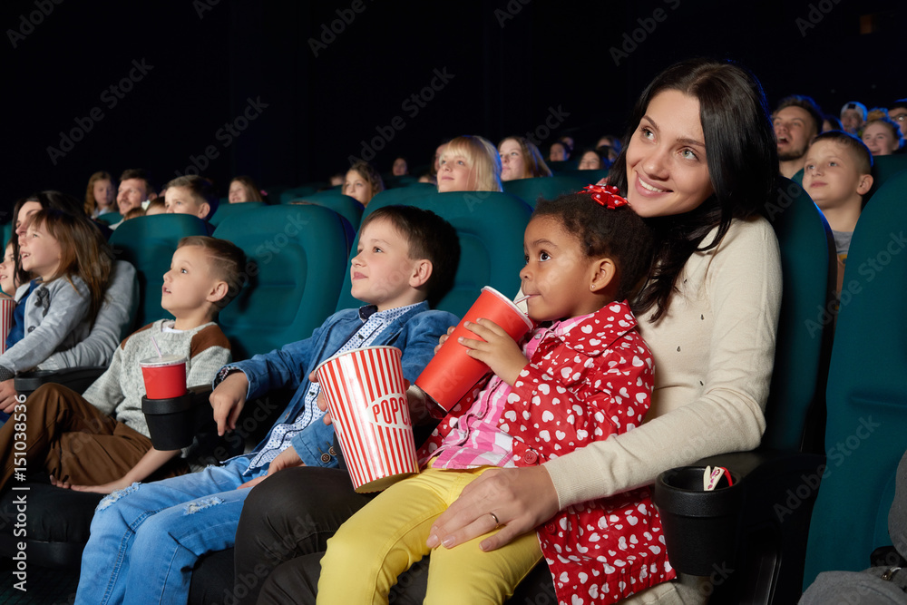 Fototapeta premium Cute little girl sipping her drink sitting on the lap of her mother at the cinema enjoying watching a cartoon copyspace family happiness positivity entertaining joyous childhood preschool.