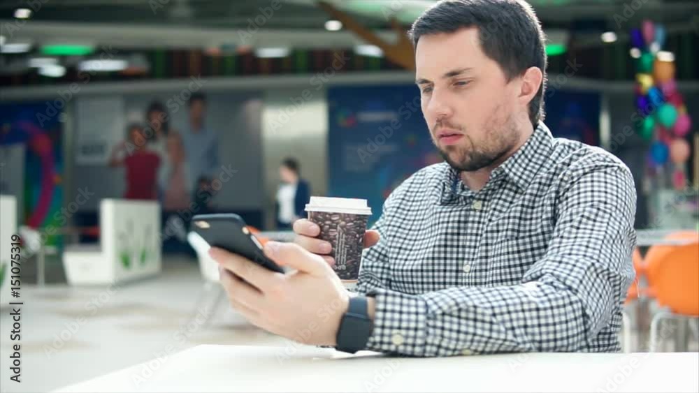 A young man with a serious appearance prints a message on his mobile phone, in his other hand is a hot coffee in a plastic glass. Person holds a smartphone in his hand, he sits in a public place. A young man with a serious appearance prints a message on his mobile phone, in his other hand is a hot coffee in a plastic glass. Person holds a smartphone in his hand, he sits in a public place.