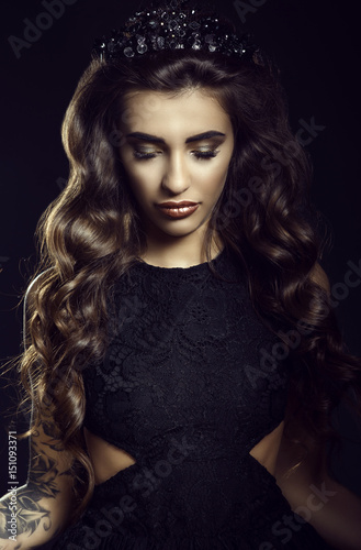 Portrait of gorgeous young model with dark long silky wavy hair, thick eyelashes and full lips wearing lace dress and jewel crown standing on black background with her eyes down. Studio shot