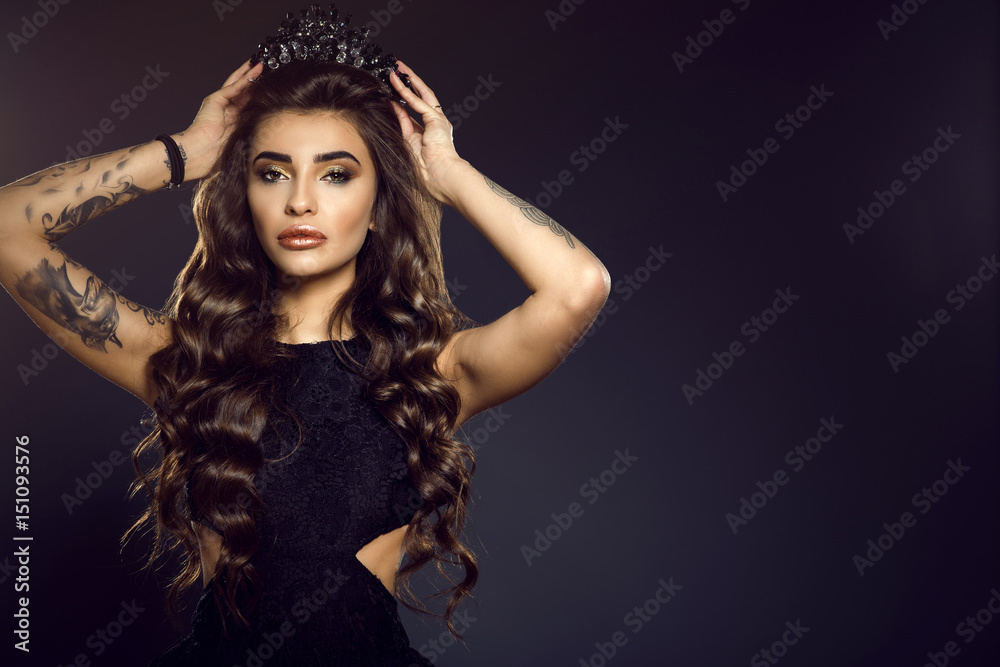 Portrait of gorgeous glam tattooed model with long wavy silky shiny hair and provocative make up putting up a jewel crown on her head. Isolated on black background with backlight. Copy-space Portrait of gorgeous glam tattooed model with long wavy silky shiny hair and provocative make up putting up a jewel crown on her head. Isolated on black background with backlight. Copy-space
