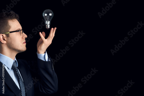 A man in a business suit looks at a lightbulb on a black background. A businessman holds a lightbulb in the air as a symbol of knowledge and ideas. © drouk