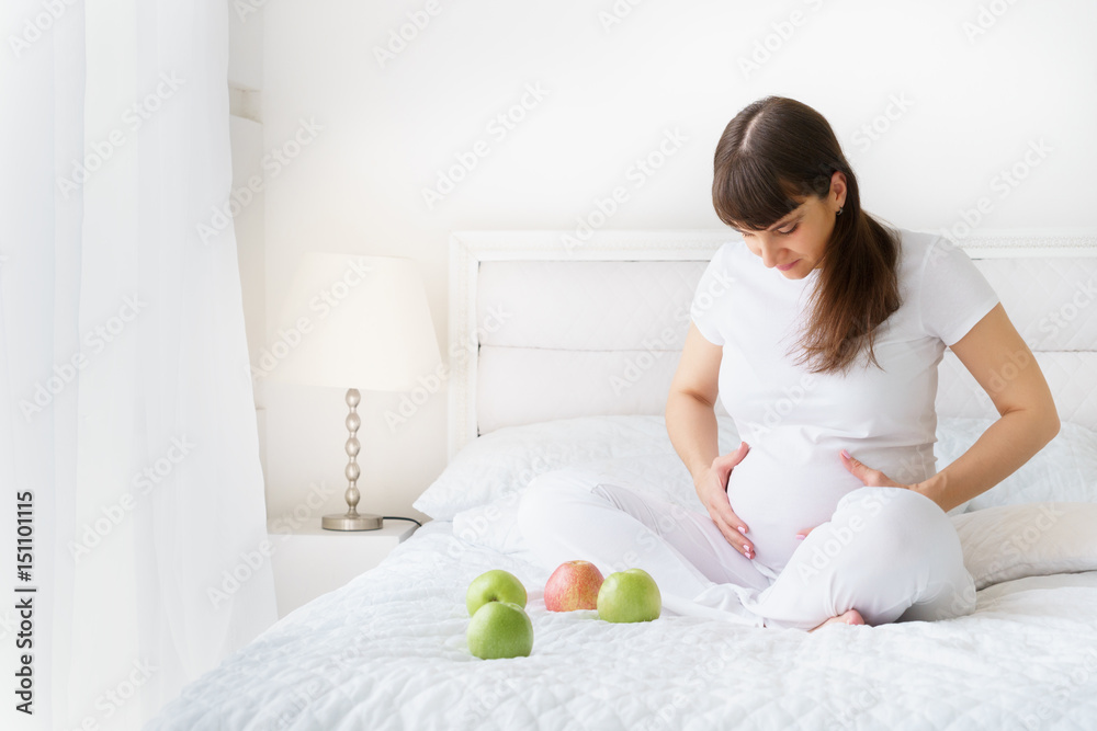 One european pregnant wooman is sitting on white bed with three green apples and smilng touching her abdomen in bedroom One european pregnant wooman is sitting on white bed with three green apples and smilng touching her abdomen in bedroom