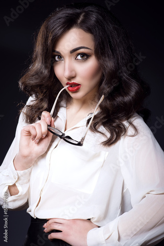 Beautiful young business woman with dark wavy hair and red lips wearing white silk blouse keeping the side of glasses in her mouth. Isolated on black background. Studio shot