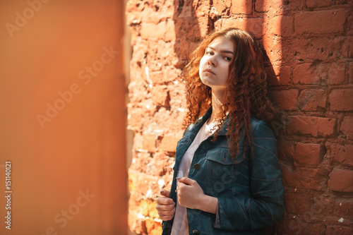 Outdoor portrait of a beautiful young white girl with red curly hair and long legs posing in a vintage old red brick yard. Lifestyle.