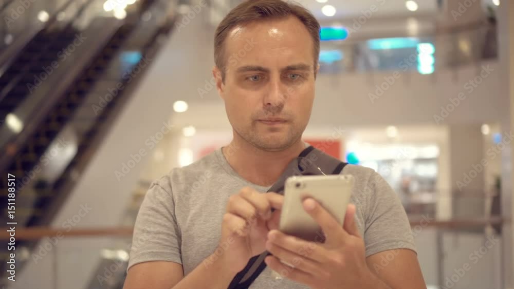 Young guy standing in shopping mall using smartphone, browsing, reading news, chatting with friends