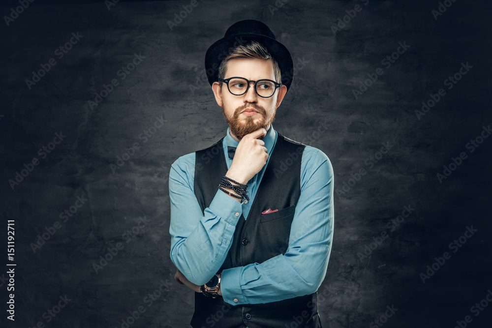 An elegant bearded hipster male dressed in a blue shirt, waistcoat and cylinder hat over grey vignette background.