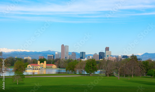 Downtown Denver skyline buildings, on a bright clear summer morning with lake and trees in foreground and snowcapped mountains in background.