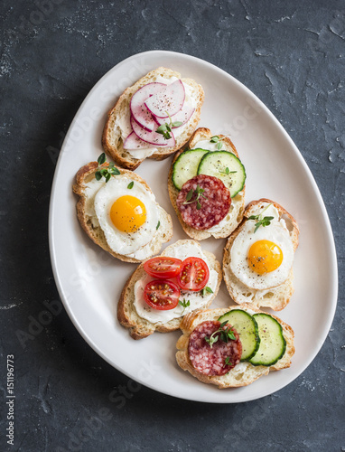 Variety of mini sandwiches with cream cheese, vegetables, quail eggs and salami. Sandwiches with cheese, cucumber, radish, tomatoes, salami, quail eggs on a dark background, top view